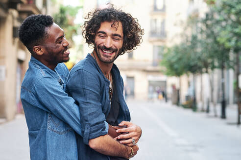Happy gay man embracing male friend from behind on road - AGOF00140