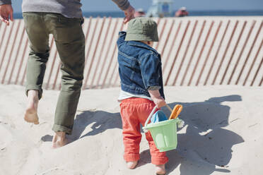 Vater hält die Hände eines Jungen, der einen Spielzeugeimer trägt, während er am Strand spazieren geht - ACTF00040