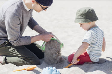 Mann und Sohn spielen mit Sand zusammen am Strand während eines sonnigen Tages - ACTF00046