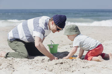 Lächelnder Mann gräbt mit seinem Sohn am Strand in der Sonne im Sand - ACTF00060