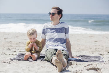 Entspannter Mann, der wegschaut, während er neben seinem Sohn sitzt und einen Snack am Strand isst - ACTF00068