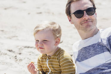 Smiling man wearing sunglasses sitting with son at beach - ACTF00071