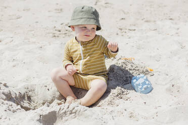 Cute little boy wearing hat sitting on sad at beach during sunny day - ACTF00075