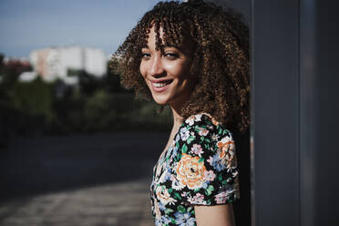 Beautiful smiling woman with curly hair standing by wall - EBBF03807