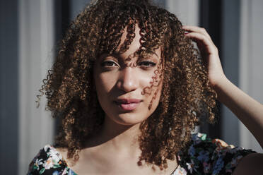 Young curly haired woman standing with hand in hair during sunny day - EBBF03808