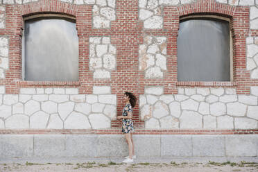 Young woman standing by brick wall looking away - EBBF03815