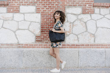 Smiling beautiful woman carrying purse while standing by brick wall - EBBF03816