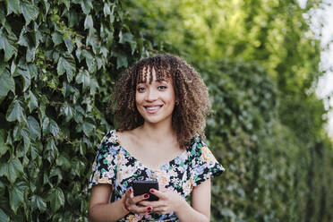 Smiling young woman holding smart phone while standing by ivy plant - EBBF03817