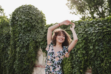 Happy young woman with arms raised dancing at garden - EBBF03818