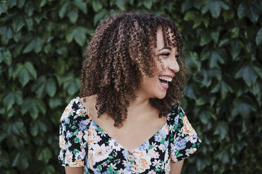 Cheerful young woman with curly hair standing in front of ivy plant - EBBF03819