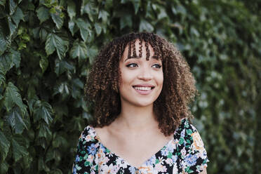 Beautiful woman with curly hair looking away while standing by ivy plant - EBBF03820