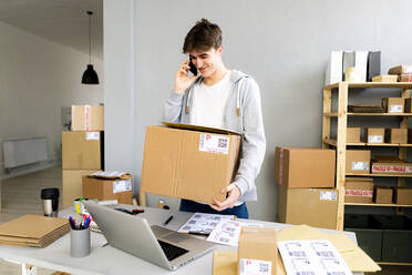 Lächelnde männliche Fachkraft, die mit einem Smartphone spricht, während sie eine Pappschachtel im Büro trägt - GIOF12767