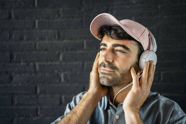 Young man looking away while listening music through headphones in front of wall - RCPF01126