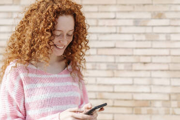 Fröhliche rothaarige Frau, die mit ihrem Smartphone vor einer Wand Textnachrichten verschickt - JCZF00716