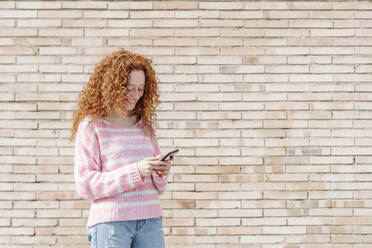 Junge Frau mit lockigem Haar, die ein Mobiltelefon benutzt, während sie vor einer Mauer steht - JCZF00717