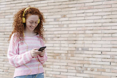 Lächelnde junge Frau, die ein Smartphone benutzt, während sie über Kopfhörer an der Wand Musik hört - JCZF00718