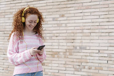 Smiling young woman using smart phone while listening music through headphones by wall - JCZF00718