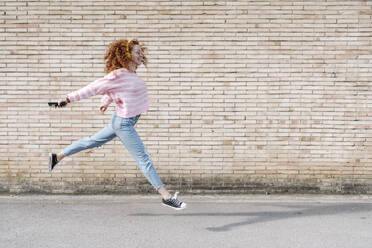 Junge rothaarige Frau mit ausgestreckten Armen springt beim Hören von Musik an der Wand - JCZF00722