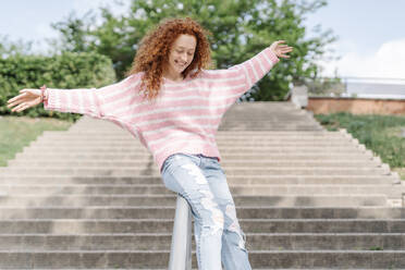 Happy redhead woman with arms outstretched sliding down on railing at park - JCZF00724