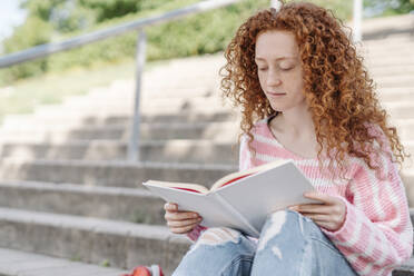 Rothaarige Frau mit lockigem Haar liest ein Buch, während sie auf einer Treppe sitzt - JCZF00725