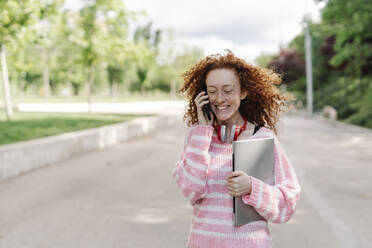 Fröhliche junge Frau mit Laptop, die im Park mit ihrem Handy telefoniert - JCZF00730