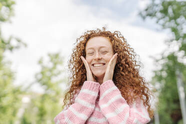 Fröhliche rothaarige Frau mit geschlossenen Augen, die ihre Wangen berührt, während sie im Park steht - JCZF00735
