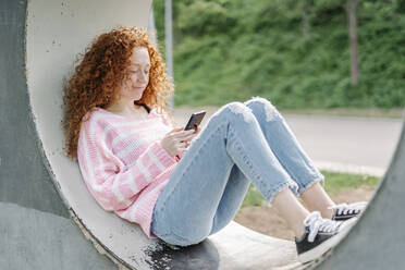 Rothaarige Frau mit lockigem Haar, die im Park liegend mit ihrem Handy im Internet surft - JCZF00738