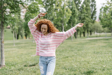 Cheerful young redhead woman dancing with arms raised at park - JCZF00742