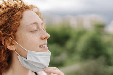 Young woman with eyes closed removing protective face mask during pandemic - JCZF00752