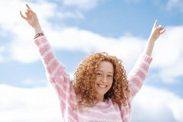 Cheerful redhead woman with arms raised standing under sky - JCZF00753