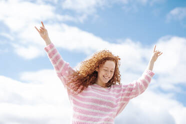 Cheerful young curly haired woman with arms raised standing under sky - JCZF00754
