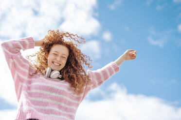 Cheerful redhead woman wearing headphones dancing under sky - JCZF00757