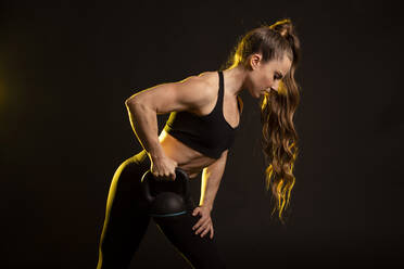 Aktive Frau, die beim Üben eine Kettlebell aufhebt, vor schwarzem Hintergrund - GMLF01199