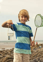 Blond boy holding jar while fishing at beach - AJOF01419