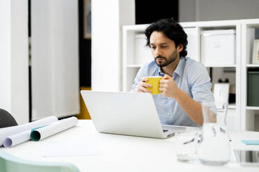 Männlicher Unternehmer mit Kaffeetasse und Laptop am Schreibtisch - GIOF12818