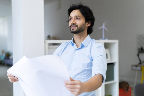 Thoughtful businessman holding paper while standing in office - GIOF12826