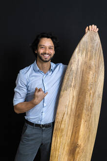 Smiling businessman with Shaka sign holding surfboard standing against black background - GIOF12829