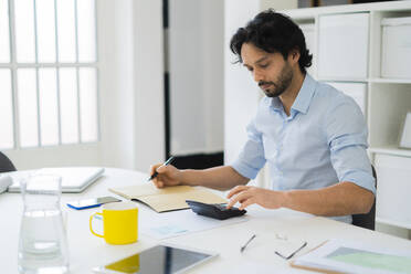 Junger männlicher Unternehmer, der bei der Arbeit im Büro einen Taschenrechner benutzt - GIOF12846