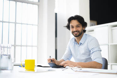 Männlicher Unternehmer, der am Schreibtisch sitzt und lächelt - GIOF12849