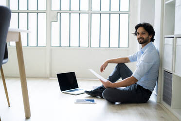 Male entrepreneur with digital tablet and laptop sitting on floor in office - GIOF12856