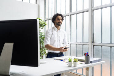 Männlicher Unternehmer mit Mobiltelefon, der wegschaut, während er im Büro steht - GIOF12862