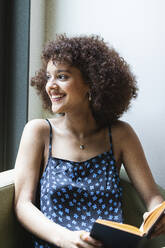 Smiling university student with book looking away while sitting in library - PNAF01791