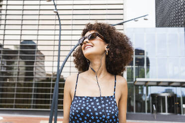 Young woman with sunglasses and in-ear headphones smiling while looking away - PNAF01810