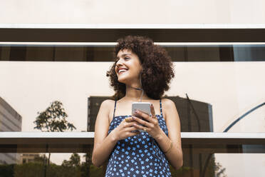 Young woman with in-ear headphones and mobile phone looking away - PNAF01812