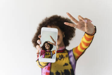 Lächelnde Frau gestikuliert bei der Aufnahme von Selfie durch Handy gegen weißen Hintergrund - PNAF01820