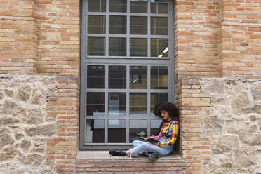 Lächelnde Frau, die ein Buch liest, während sie auf der Fensterbank der Universität sitzt - PNAF01825