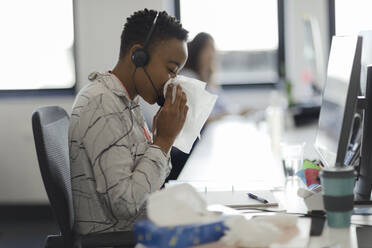Geschäftsfrau mit Headset schnäuzt Nase in Taschentuch am Bürotisch - CAIF30501