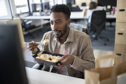 Geschäftsmann isst Sushi zum Mitnehmen am Computer im Büro - CAIF30504