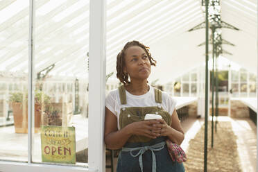 Serene garden shop owner with coffee in greenhouse doorway - CAIF30551