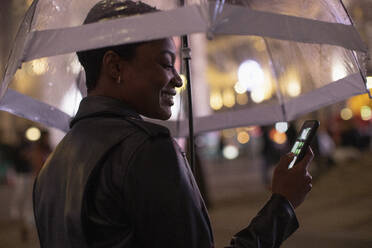 Glückliche junge Frau, die nachts unter einem Regenschirm ihr Smartphone benutzt - CAIF30630
